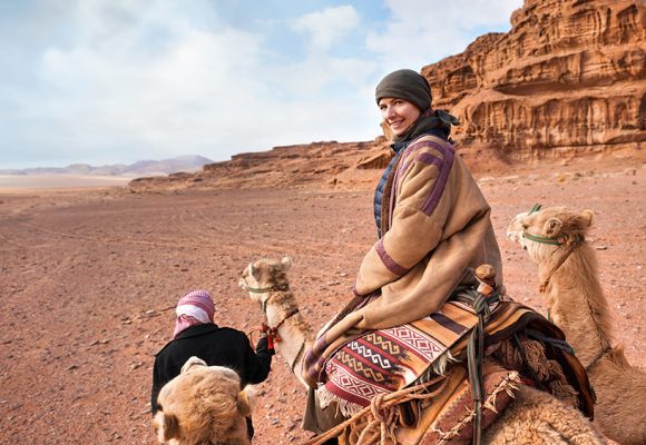 Young woman riding camel in Wadi Rum desert, looking back over her shoulder, smiling. It's quite cold so she is wearing traditional Bedouin coat - bisht - and head scarf, mountains far background